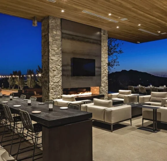 phoenix-clubhouse-1 Outdoor lounge area at dusk with a stone fireplace and mounted TV, white couches, and dark dining tables under a wooden ceiling with string lights