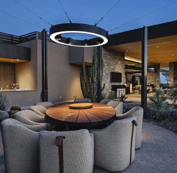 phoenix-clubhouse-2 Outdoor circular wooden table surrounded by curved beige chairs under a glowing ring chandelier at twilight.