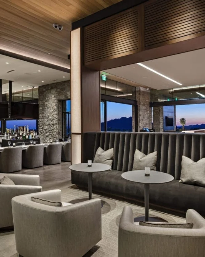 phoenix-clubhouse-3 Elegant hotel lounge with gray upholstered chairs, round tables, and a long cushioned back seat, stone walls, and large windows showing a twilight mountain view.